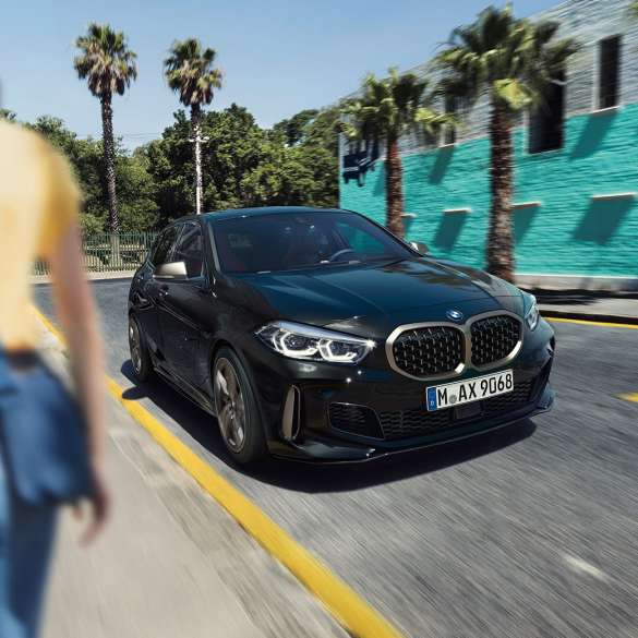 BMW M135i xDrive, perspetiva frontal a três quartos BMW M135i xDrive na pintura preto Sapphire metalizada, em movimento, perspetiva frontal a três quartos, com uma mulher em primeiro plano.