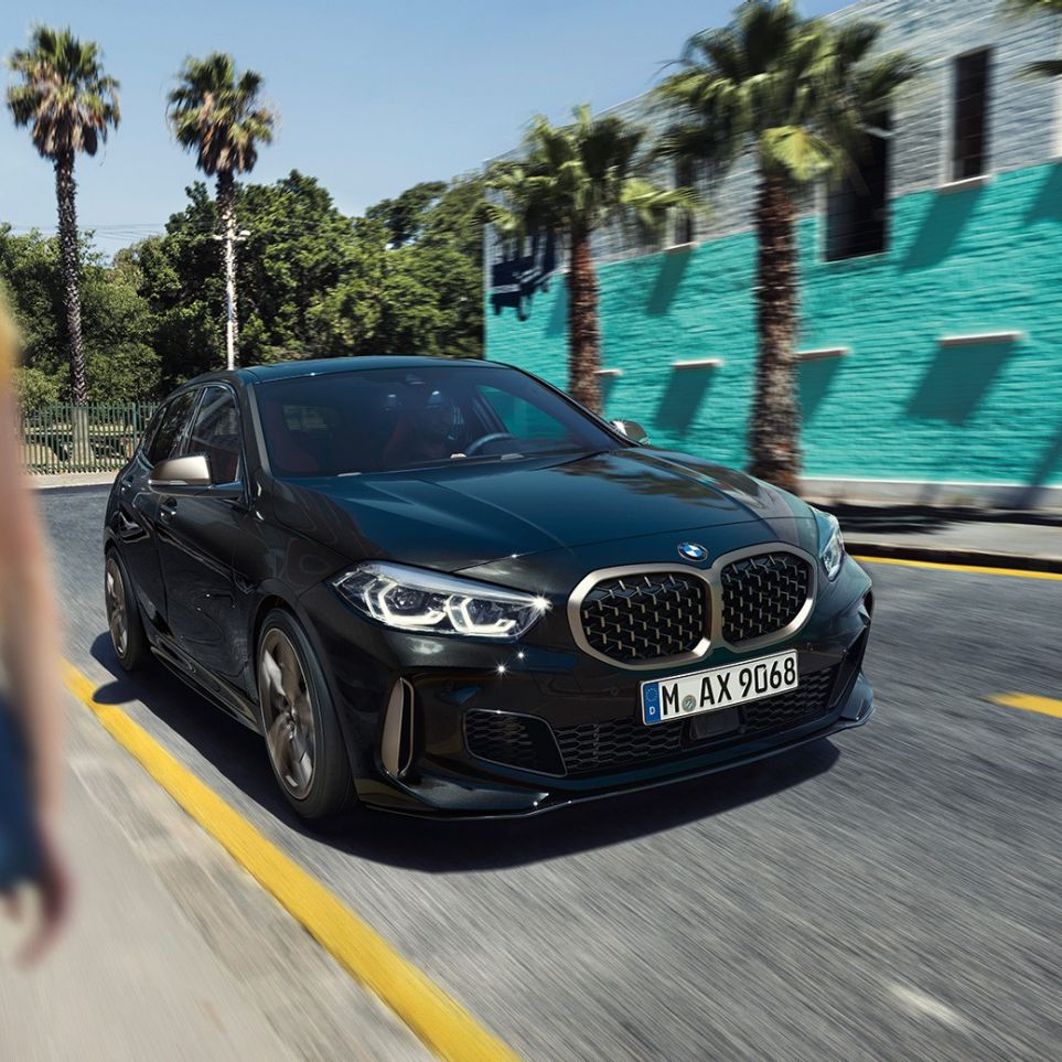 BMW M135i xDrive, perspetiva frontal a três quartos. BMW M135i xDrive na pintura preto Sapphire metalizada, em movimento, perspetiva frontal a três quartos, com uma mulher em primeiro plano.