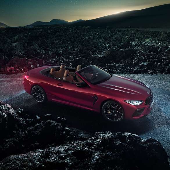 BMW M8 Competition Cabrio, perspetiva lateral a três quartos. BMW M8 Competition Cabrio na pintura vermelho Motegi metalizada com capota flexível aberta, no asfalto à noite, perspetiva lateral a três quartos.