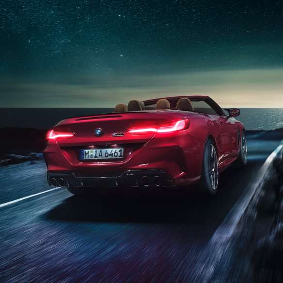 BMW M8 Competition Cabrio, perspetiva traseira a três quartos. BMW M8 Competition Cabrio na pintura vermelho Motegi metalizada com capota flexível aberta, em movimento à noite numa estrada junto à costa, perspetiva traseira a três quartos.