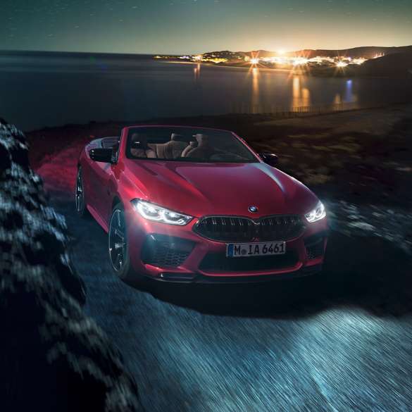 BMW M8 Competition Cabrio, perspetiva frontal a três quartos. BMW M8 Competition Cabrio na pintura vermelho Motegi metalizada com capota flexível aberta, em movimento à noite numa estrada junto à costa, perspetiva frontal a três quartos.