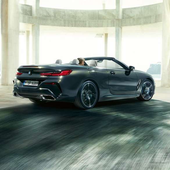 BMW M850i xDrive Cabrio, perspetiva traseira a três quartos. BMW M850i xDrive Cabrio na pintura cinza Dravit metalizada com capota flexível aberta, a passar por um edifício em betão, perspetiva traseira a três quartos.