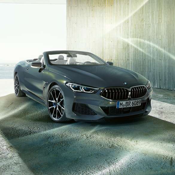 BMW M850i xDrive Cabrio, perspetiva frontal a três quartos. BMW M850i xDrive Cabrio na pintura cinza Dravit metalizada com capota flexível aberta, em frente ao mar, perspetiva frontal a três quartos.