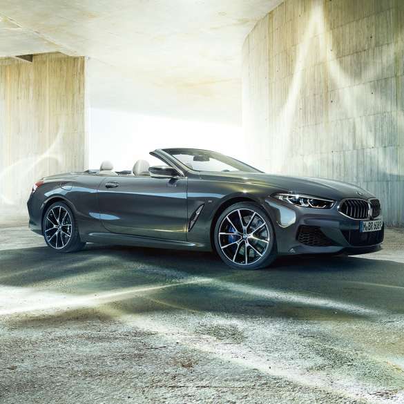 BMW M850i xDrive Cabrio, perspetiva lateral a três quartos. BMW M850i xDrive Cabrio na pintura cinza Dravit metalizada com capota flexível aberta, em frente a um edifício em betão, perspetiva lateral a três quartos.