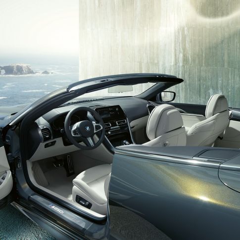 BMW M850i xDrive Cabrio, interior. BMW M850i xDrive Cabrio em cinza Dravit metalizada com capota flexível aberta, interior com volante M em pele e estofos em pele Merino BMW Individual em branco Ivory.