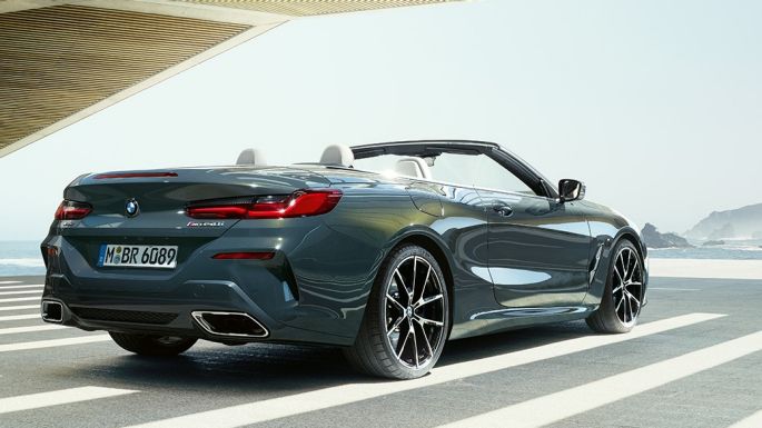 BMW M850i xDrive Cabrio, perspetiva traseira a três quartos. BMW M850i xDrive Cabrio na pintura cinza Dravit metalizada com capota flexível aberta, a estacionar num local com perspetiva para o mar, perspetiva traseira a três quartos.