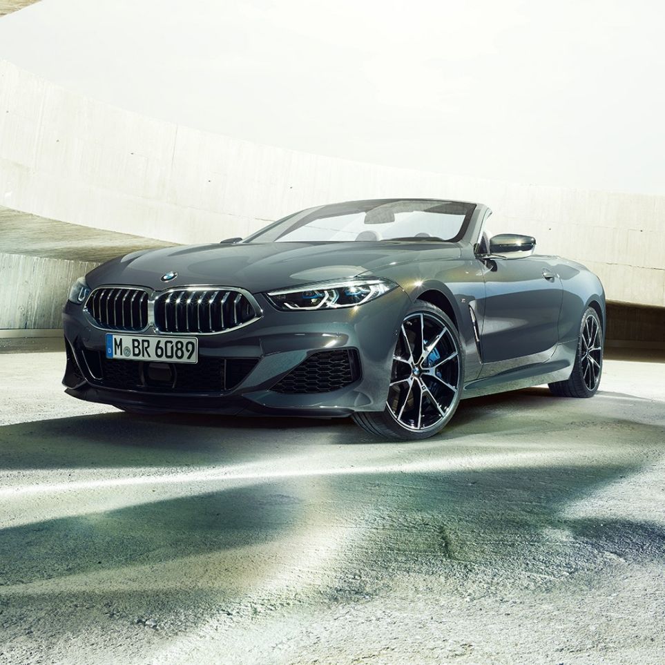 BMW M850i xDrive Cabrio, perspetiva frontal a três quartos. BMW M850i xDrive Cabrio na pintura cinza Dravit metalizada com capota flexível aberta, parado no interior de um edifício em betão, perspetiva frontal a três quartos.