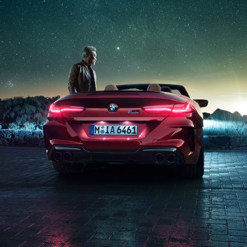 BMW M8 Competition Cabrio, perspetiva traseira. BMW M8 Competition Cabrio na pintura vermelho Motegi metalizada com capota flexível aberta, parado, com um homem sob um céu estrelado, perspetiva traseira.