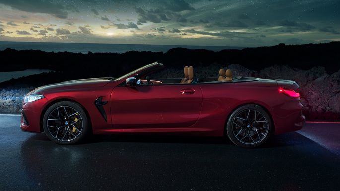 BMW M8 Competition Cabrio, perspetiva lateral. BMW M8 Competition Cabrio na pintura vermelho Motegi metalizada com capota flexível aberta, à noite, sob um céu estrelado, perspetiva lateral.