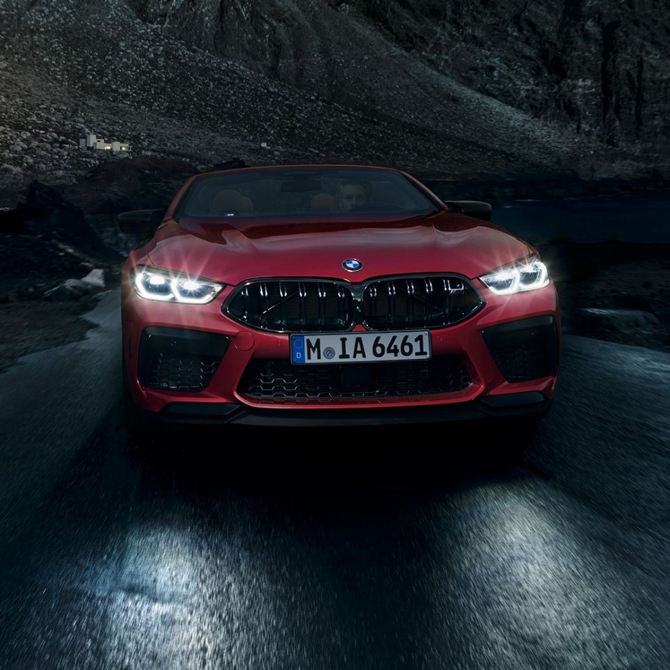 BMW M8 Competition Cabrio, perspetiva frontal. BMW M8 Competition Cabrio na pintura vermelho Motegi metalizada com faróis ligados, em movimento, à noite, perspetiva frontal.