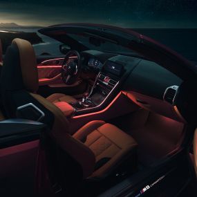BMW M8 Competition Cabrio, interior. BMW M8 Competition Cabrio com capota fexível aberta, interior com luz ambiente em vermelho, à noite.