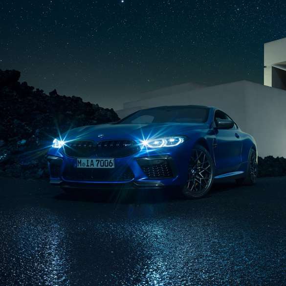 BMW M8 Competition Coupé, perspetiva frontal a três quartos. BMW M8 Competition Coupé na pintura azul Marina Bay metalizada com faróis LED iluminados, parado em frente a uma moradia, à noite, perspetiva frontal a três quartos.