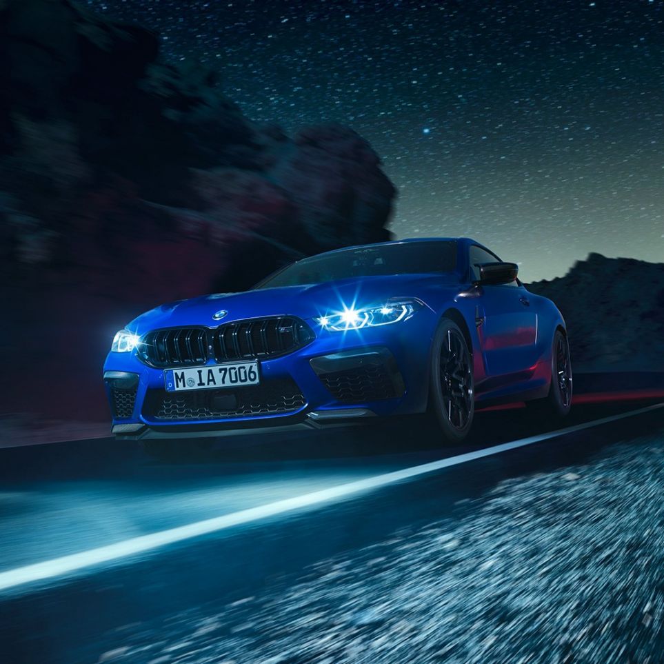 BMW M8 Competition Coupé em movimento, perspetiva frontal a três quartos. BMW M8 Competition Coupé na pintura azul Marina Bay metalizada com faróis LED iluminados, em movimento, à noite, perspetiva frontal a três quartos.