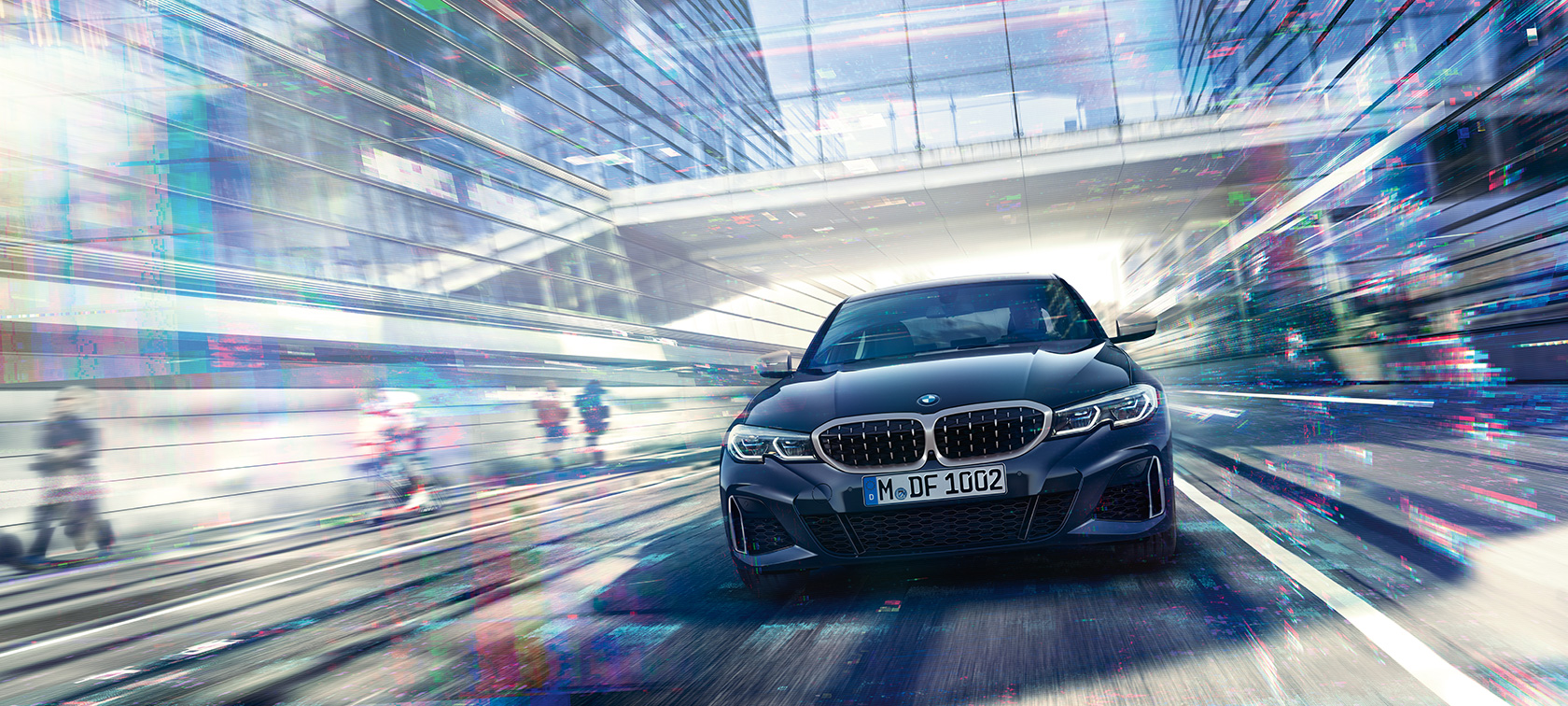 BMW Série 3 Berlina, perspetiva frontal do BMW M340i xDrive. Perspetiva frontal do BMW M340i xDrive Berlina em movimento num cenário urbano, entre edifícios em vidro.