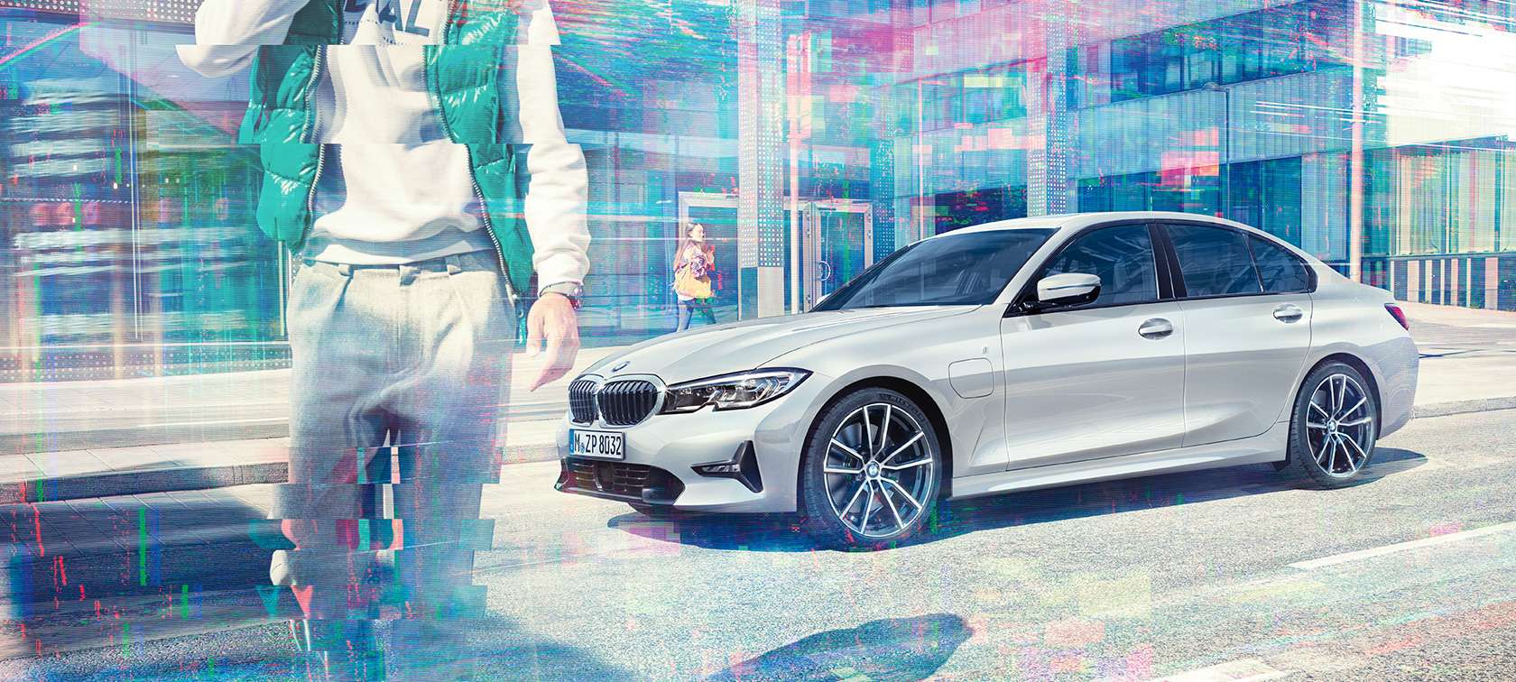 BMW Série 3 Berlina, perspetiva lateral do BMW 330e. Perspetiva lateral a três quartos do BMW 330e Berlina parado em frente a um edifício em vidro com peões em movimento.