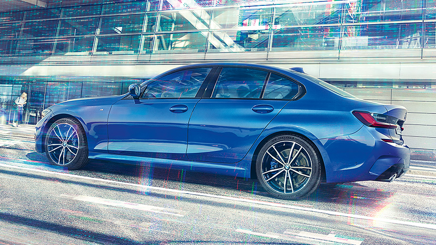 BMW Série 3 Berlina (G20) durante o dia em frente a um edifício em vidro. Perspetiva lateral do BMW Série 3 Berlina (G20) parado, pintura azul Portimao, durante o dia em frente a um edifício em vidro.