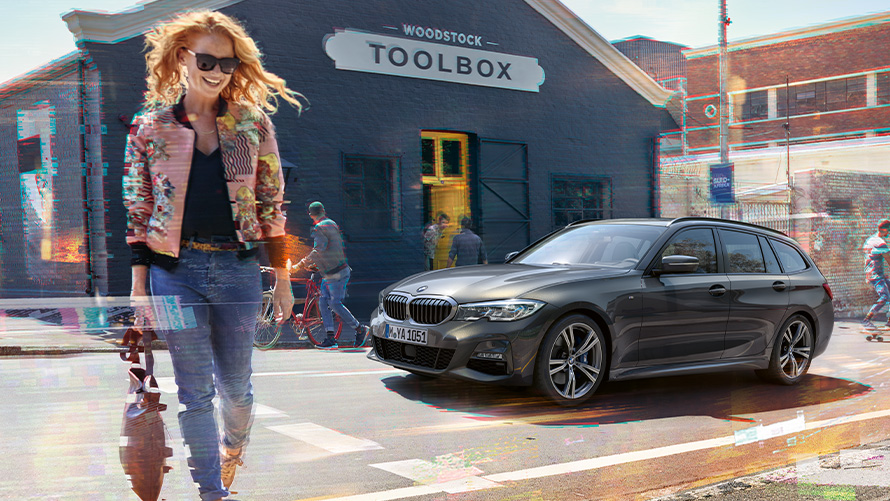 BMW Série 3 Touring (G21), perspetiva frontal a três quartos com uma mulher à frente de uma oficina de ferramentas de marcenaria. Uma mulher a atravessar a rua à frente de um BMW Série 3 Touring (G21) à frente de uma oficina de ferramentas de marcenaria.