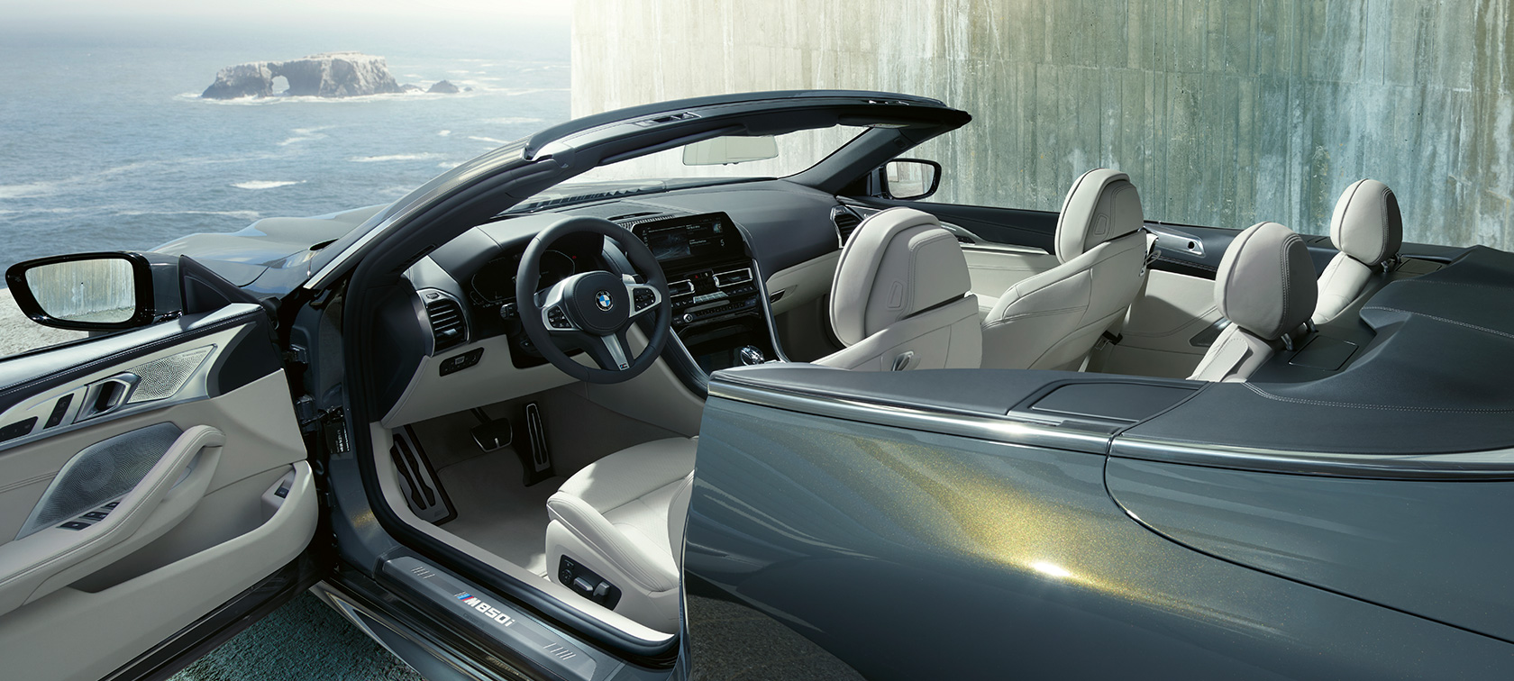 BMW M850i xDrive em cinza Dravit metalizado, perspetiva lateral, capota flexível e porta aberta. BMW M850i xDrive em cinza Dravit metalizado, perspetiva lateral, capota flexível e porta aberta.