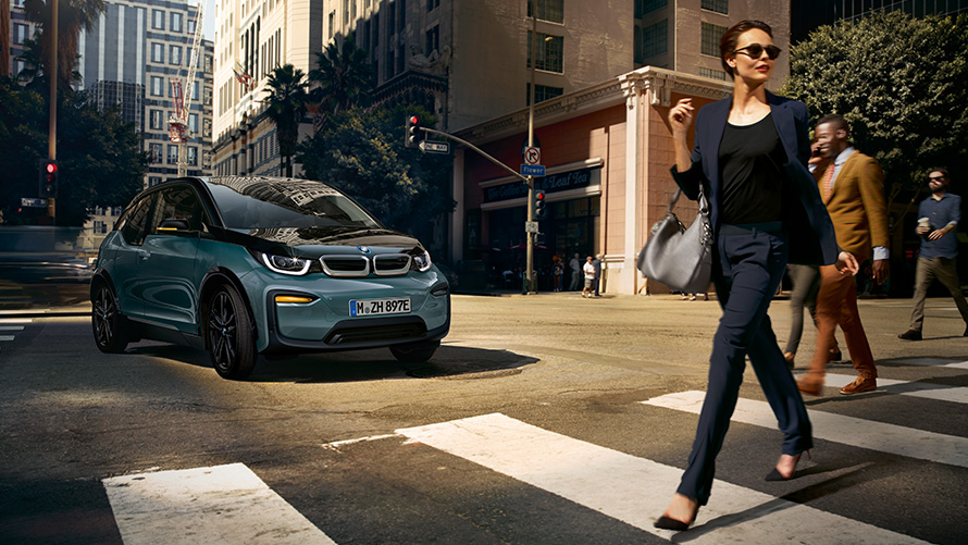 BMW i3 I01, perspetiva frontal a três quartos com mulher a atravessar uma passadeira BMW i3 I01, pintura azul Ridge Mountain metalizada com realce em cinza Frozen metalizada, perspetiva frontal a três quartos com mulher a atravessar uma passadeira