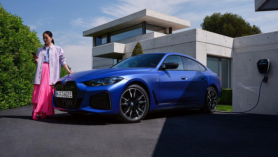 BMW i4 M50, G26, 2021, pintura azul Frozen Portimao BMW Individual metalizada, perspetiva frontal a três quartos, parado com uma mulher à frente de uma casa