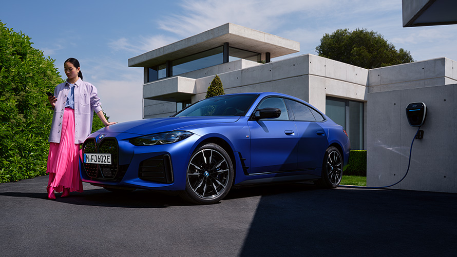 Perspetiva lateral a três quartos, BMW i4 M50, G26, 2021, com uma mulher à frente de uma casa BMW i4 M50, G26, 2021, pintura azul Frozen Portimao BMW Individual metalizada, perspetiva lateral a três quartos, com uma mulher à frente de uma casa