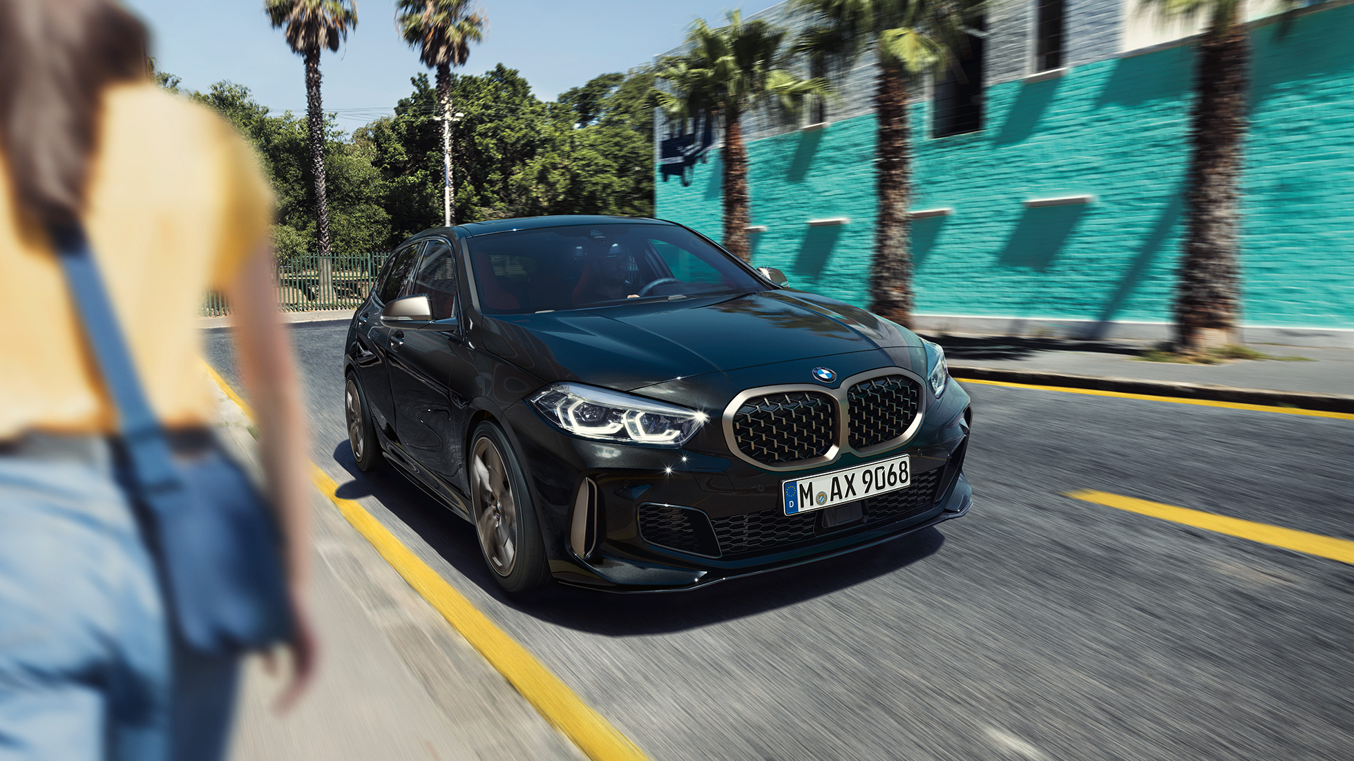 BMW M135i xDrive, perspetiva frontal a três quartos BMW M135i xDrive na pintura preto Sapphire metalizada, em movimento, perspetiva frontal a três quartos, com uma mulher em primeiro plano.