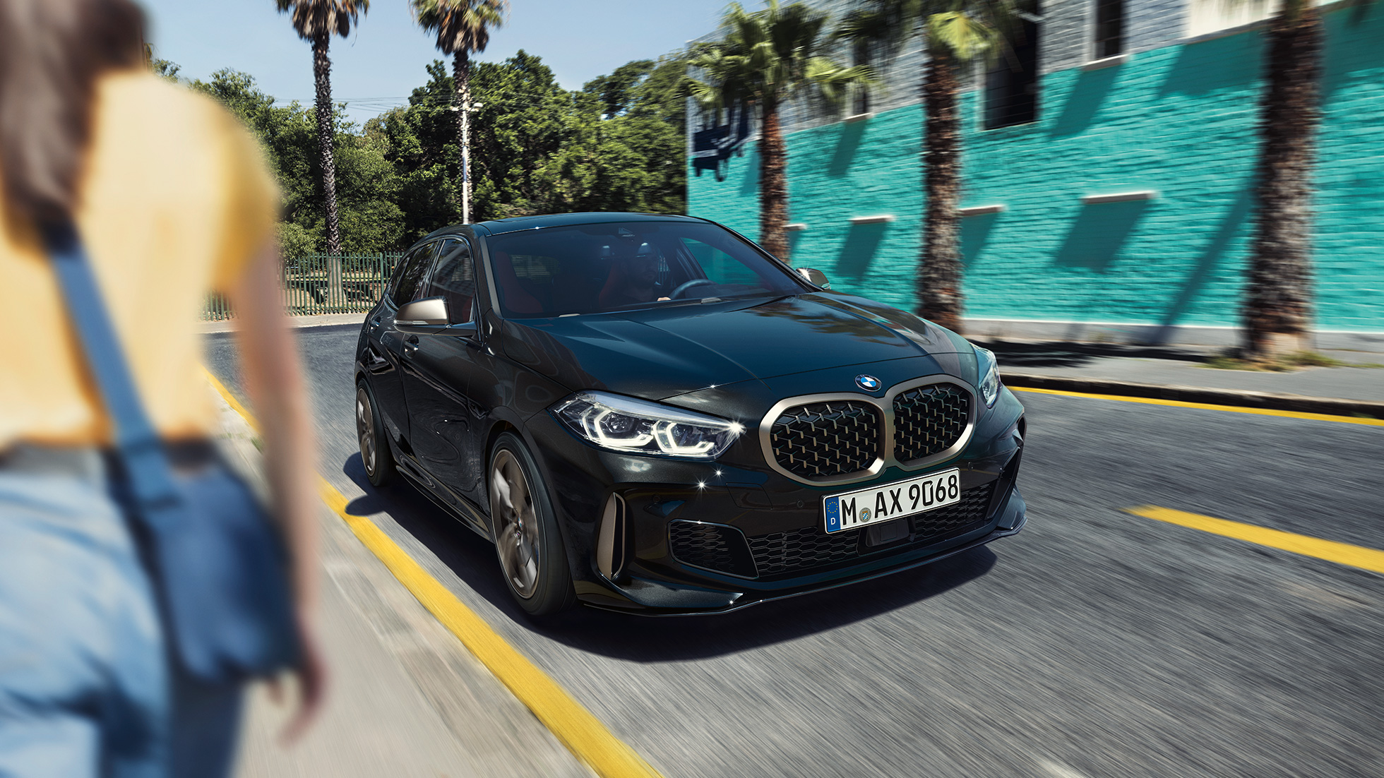 BMW M135i xDrive, perspetiva frontal a três quartos. BMW M135i xDrive na pintura preto Sapphire metalizada, em movimento, perspetiva frontal a três quartos, com uma mulher em primeiro plano.