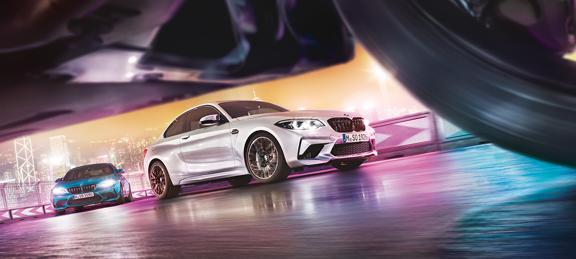 BMW M2 Competition com reflexo no asfalto. Dois BMW M2 Competition em pista, frente a uma paisagem urbana noturna, ao nível da estrada.