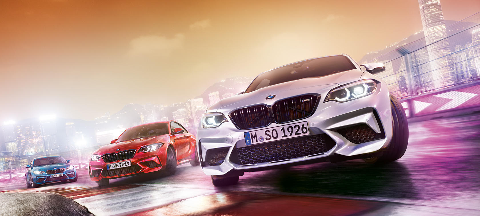 BMW M2 Competition em ação Três BMW M2 Competition em pista, frente a uma paisagem urbana noturna.
