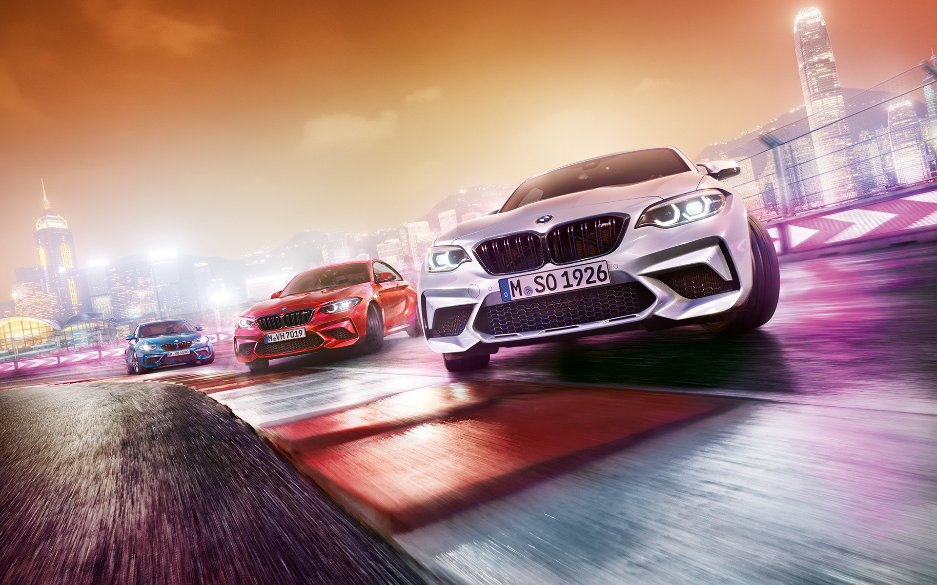 BMW M2 Competition em ação Três BMW M2 Competition em pista, frente a uma paisagem urbana noturna.
