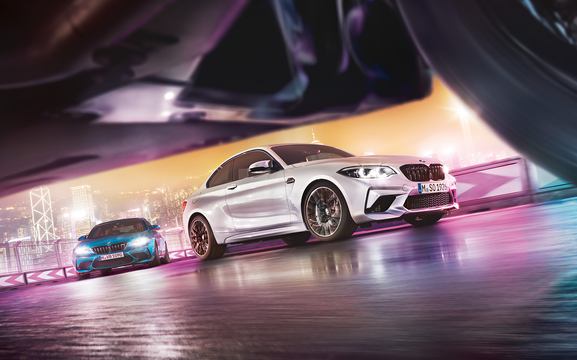 BMW M2 Competition com reflexo no asfalto. Dois BMW M2 Competition em pista, frente a uma paisagem urbana noturna, ao nível da estrada.