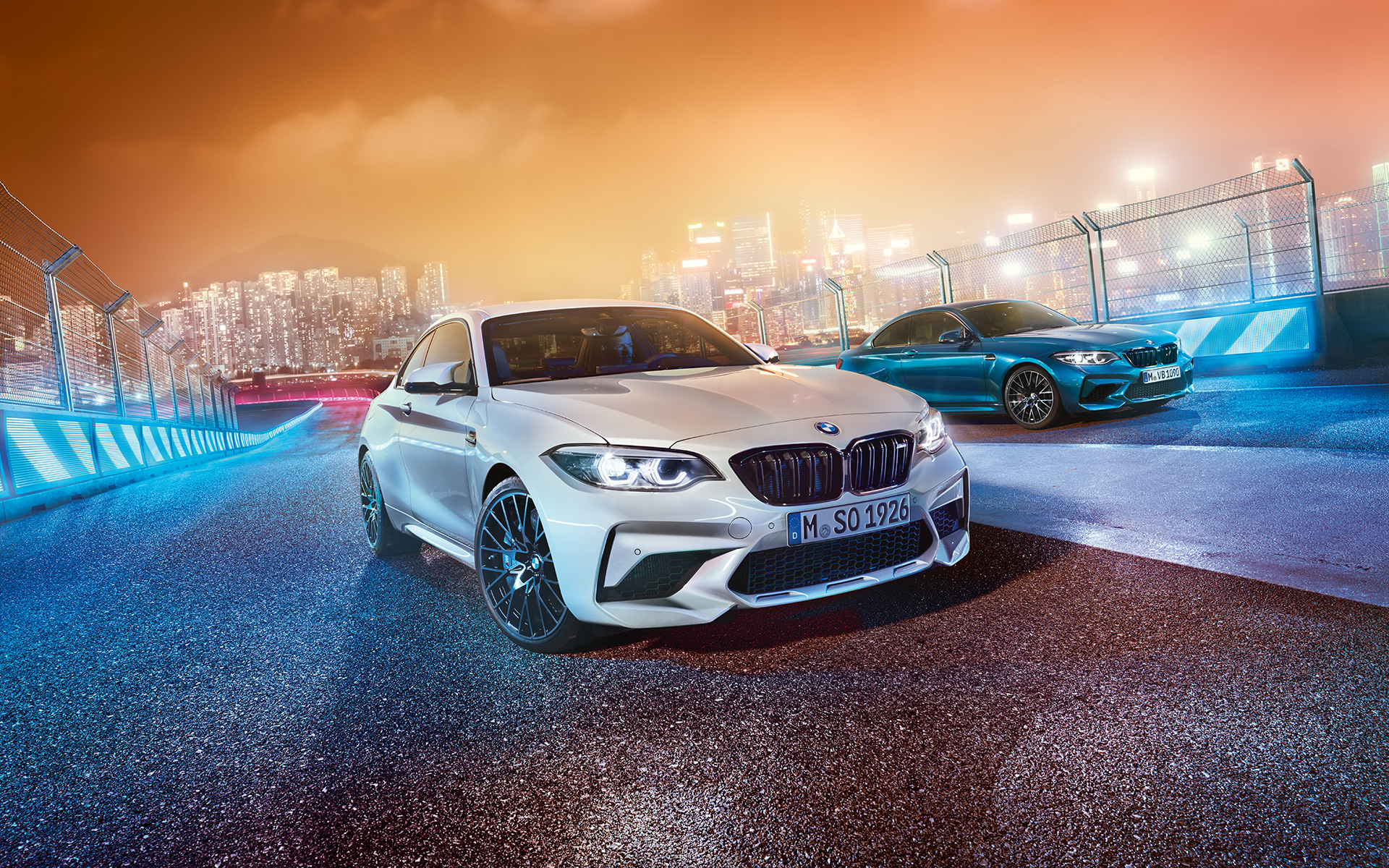 Dois BMW M2 Competition em pista Vista dianteira de dois BMW M2 Competition em branco e azul, frente a uma paisagem urbana noturna.