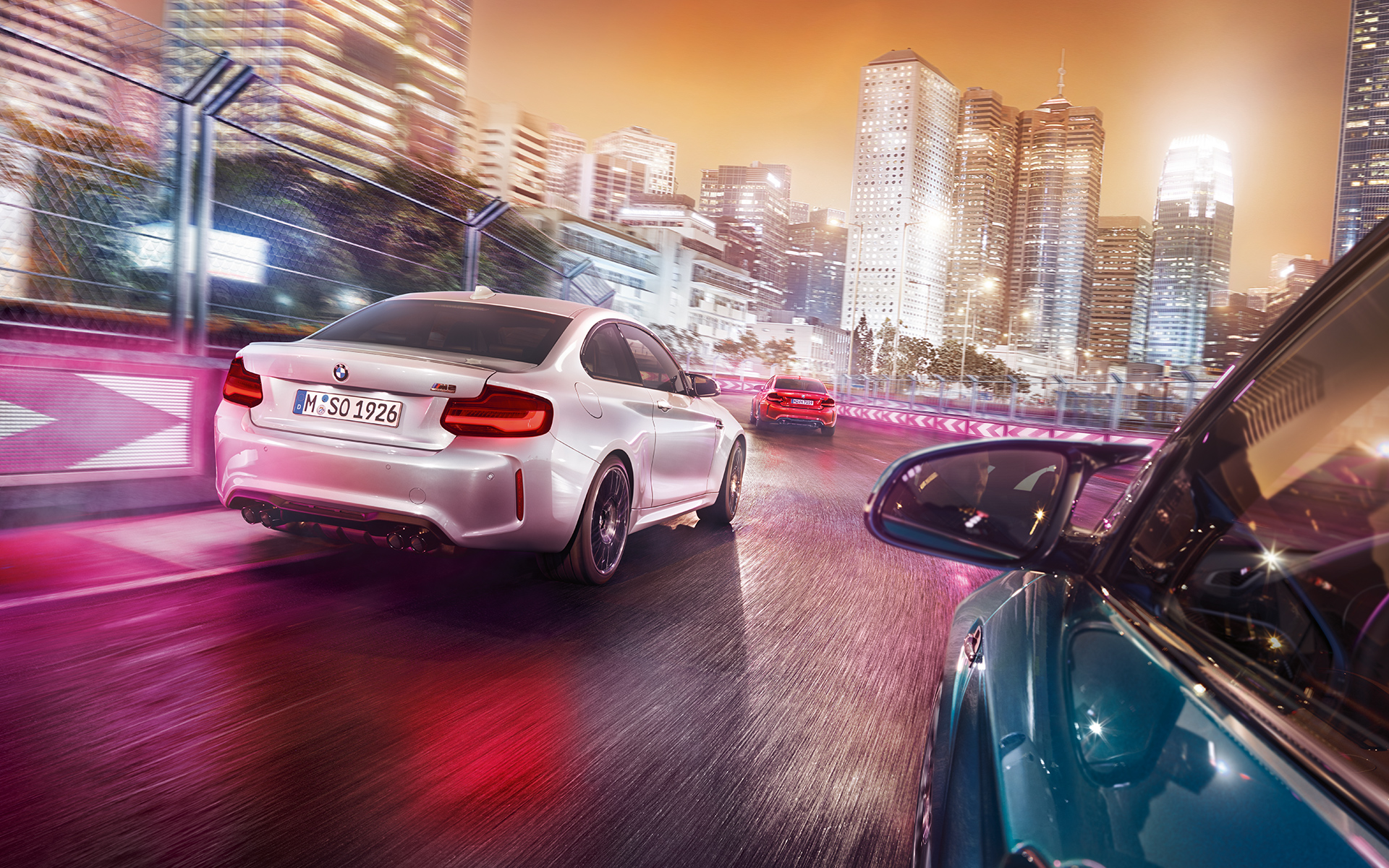 Vista traseira do BMW M2 Competition Três BMW M2 Competition em frente a uma paisagem noturna com arranha-céus.