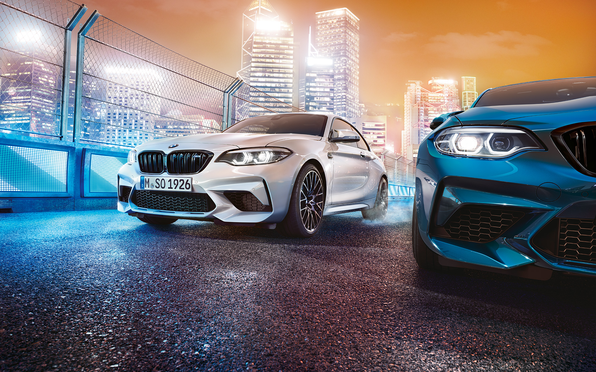 BMW M2 Competition branco e azul estacionados. Vista dianteira de dois BMW M2 Competition em branco e azul, frente a uma paisagem urbana noturna.