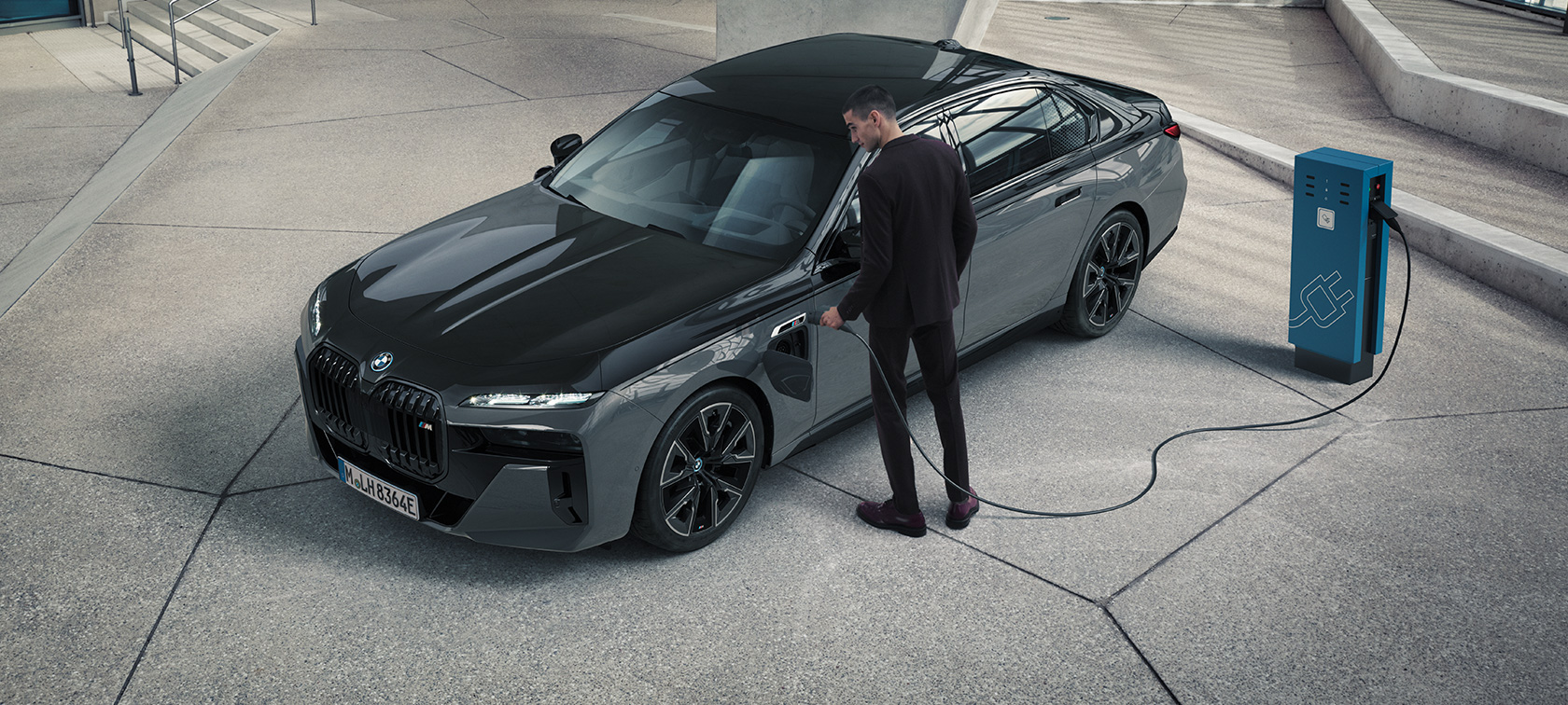 BMW M760e xDrive Berlina, G70, pack M Performance BMW M760e xDrive Berlina, G70, pack M Performance, pintura preto Sapphire BMW Individual de duas tonalidades metalizada, pintura cinza Dravite BMW Individual metalizada, perspetiva dianteira a três quartos, homem com cabo de carregamento ao lado do posto de carregamento