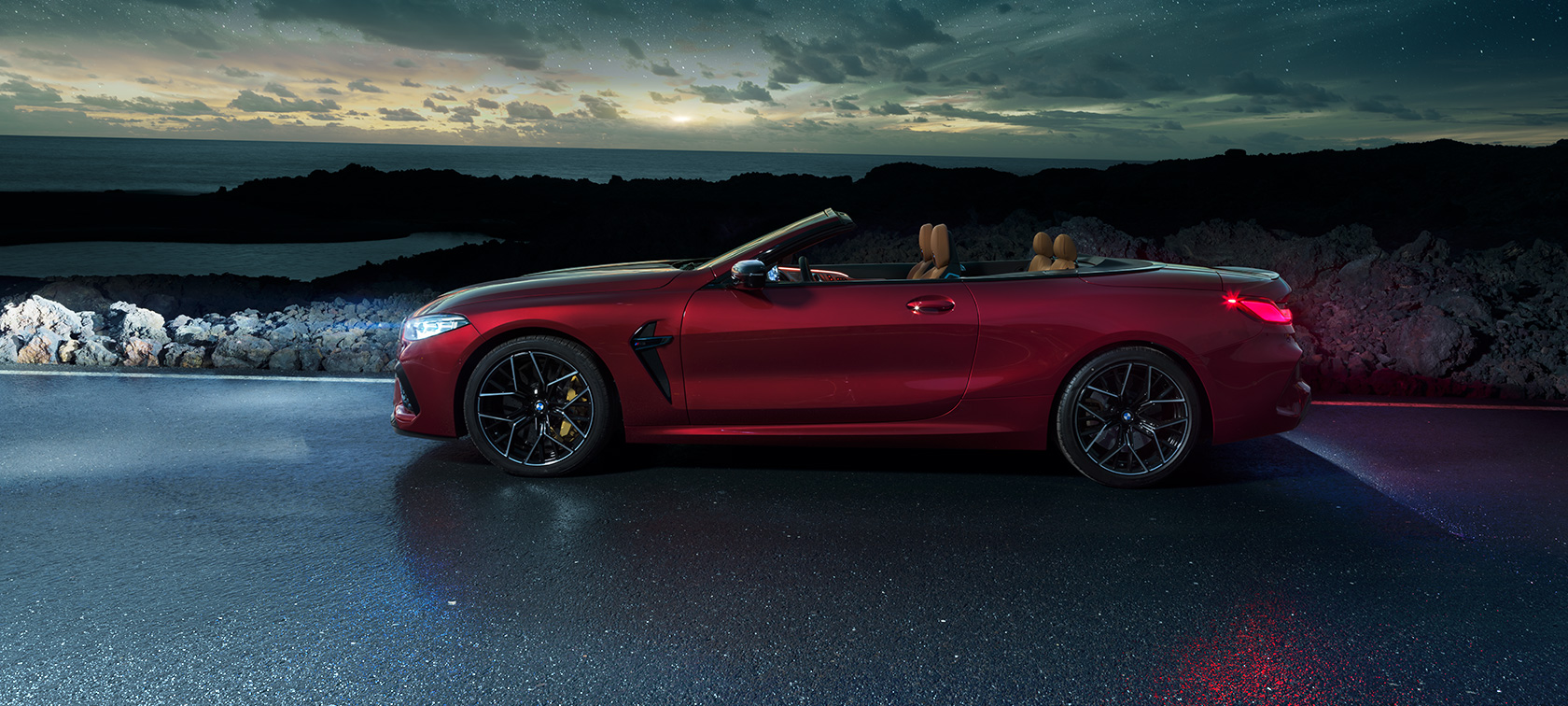 BMW M8 Competition Cabrio, perspetiva lateral. BMW M8 Competition Cabrio na pintura vermelho Motegi metalizada com capota flexível aberta, numa estrada junto à costa, sob um céu estrelado, perspetiva lateral.