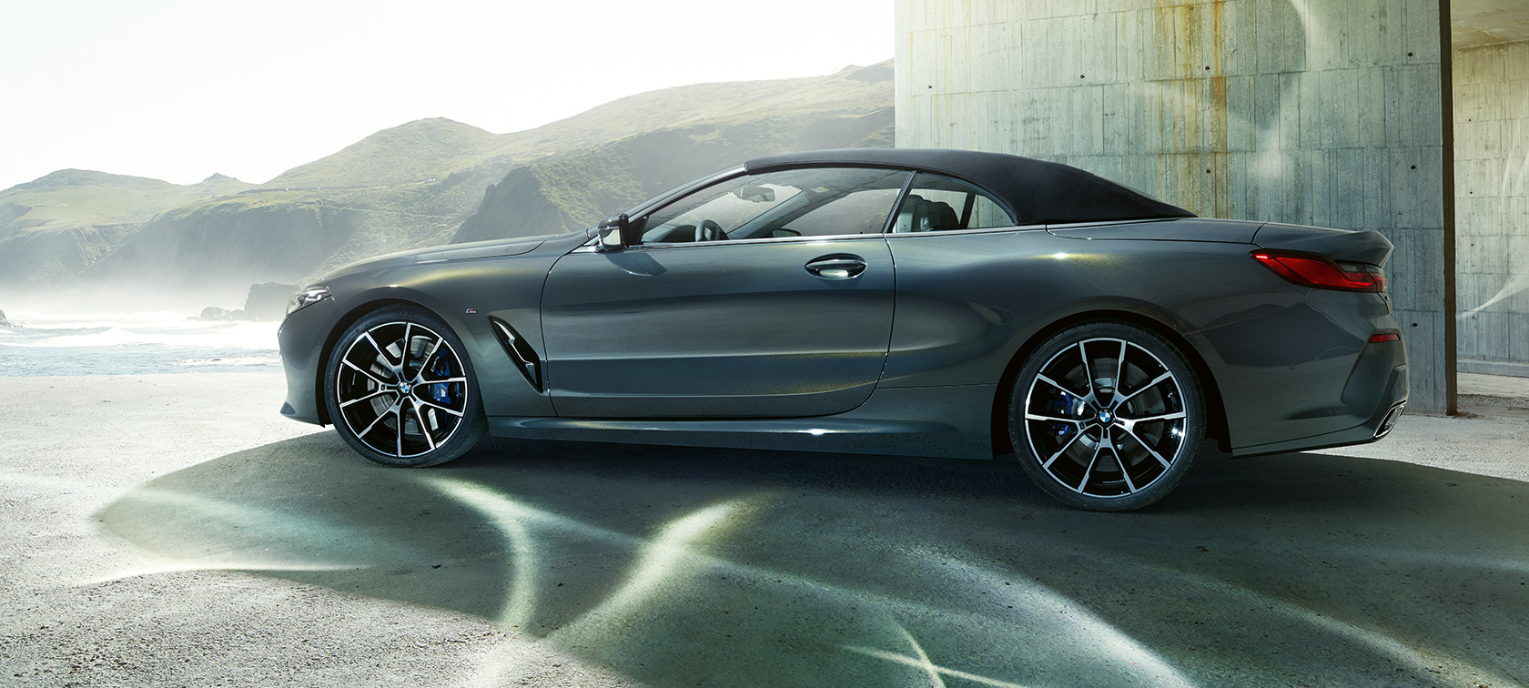 BMW M850i xDrive Cabrio, perspetiva lateral. BMW M850i xDrive Cabrio na pintura cinza Dravit metalizada com capota flexível fechada num fundo de montanhas, perspetiva lateral.