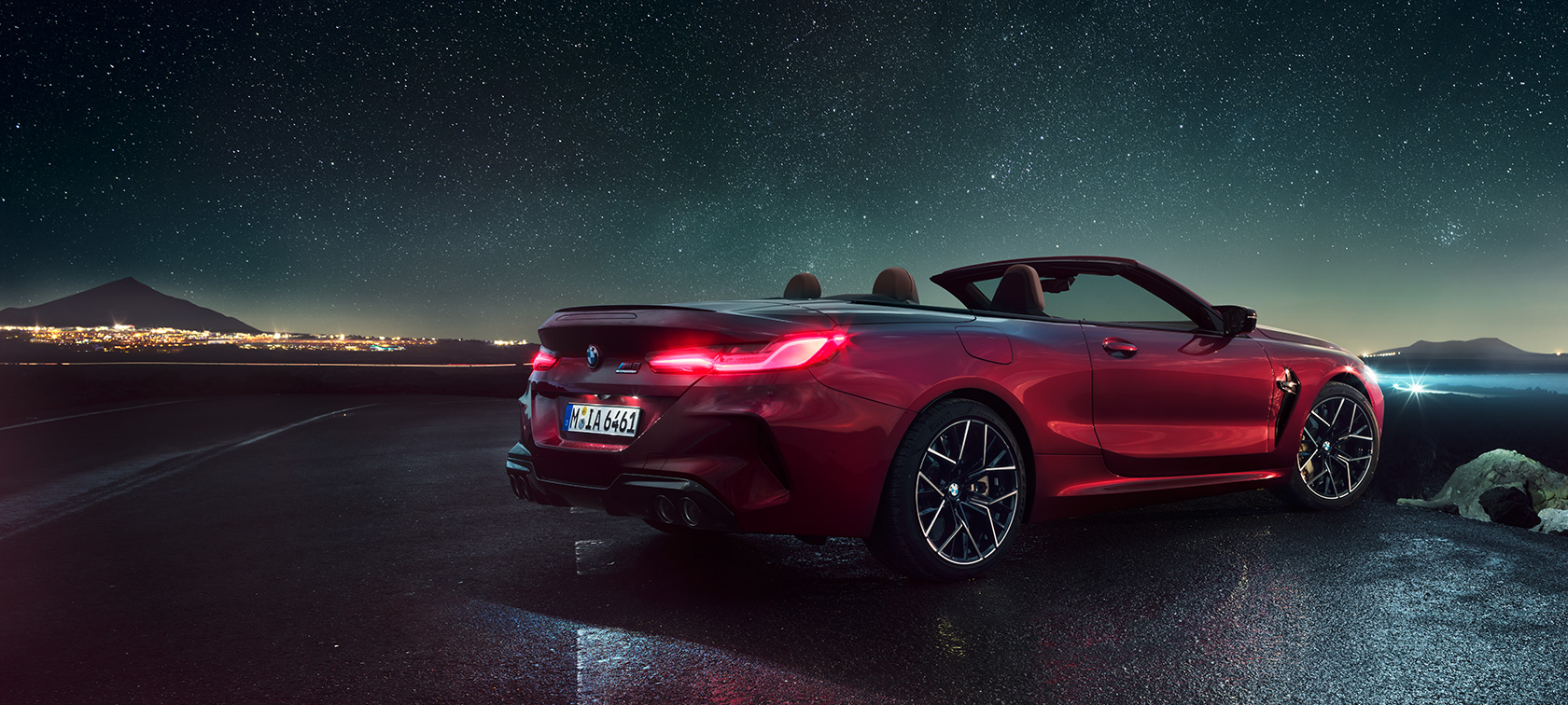 BMW M8 Competition Cabrio, perspetiva traseira a três quartos. BMW M8 Competition Cabrio na pintura vermelho Motegi metalizada com capota flexível aberta, sob um céu estrelado, perspetiva traseira a três quartos.