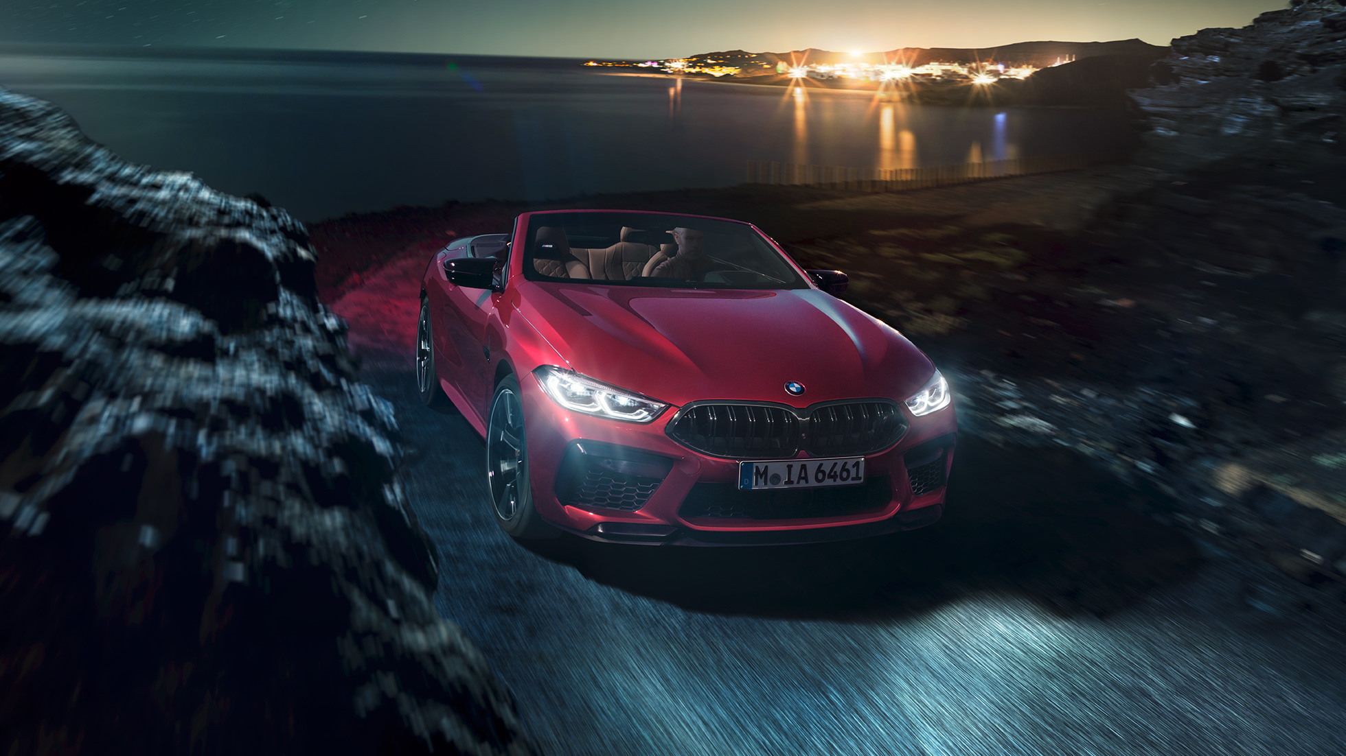 BMW M8 Competition Cabrio, vista frontal a três quartos. BMW M8 Competition Cabrio na cor exterior vermelho Motegi metalizada com a capota flexível aberta, em movimento, à noite, numa estrada junto à costa, vista frontal a três quartos.