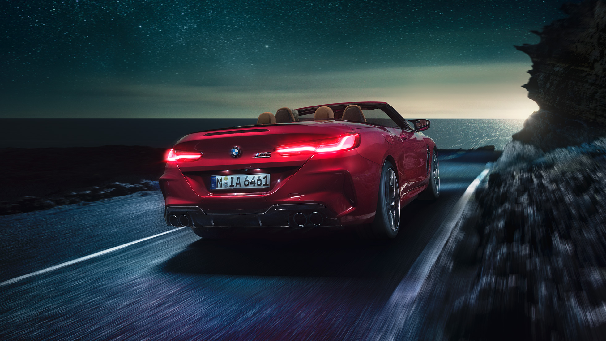 BMW M8 Competition Cabrio, vista traseira a três quartos. BMW M8 Competition Cabrio na cor exterior vermelho Motegi metalizada com a capota flexível aberta, em movimento, à noite, numa estrada junto à costa, vista traseira a três quartos.