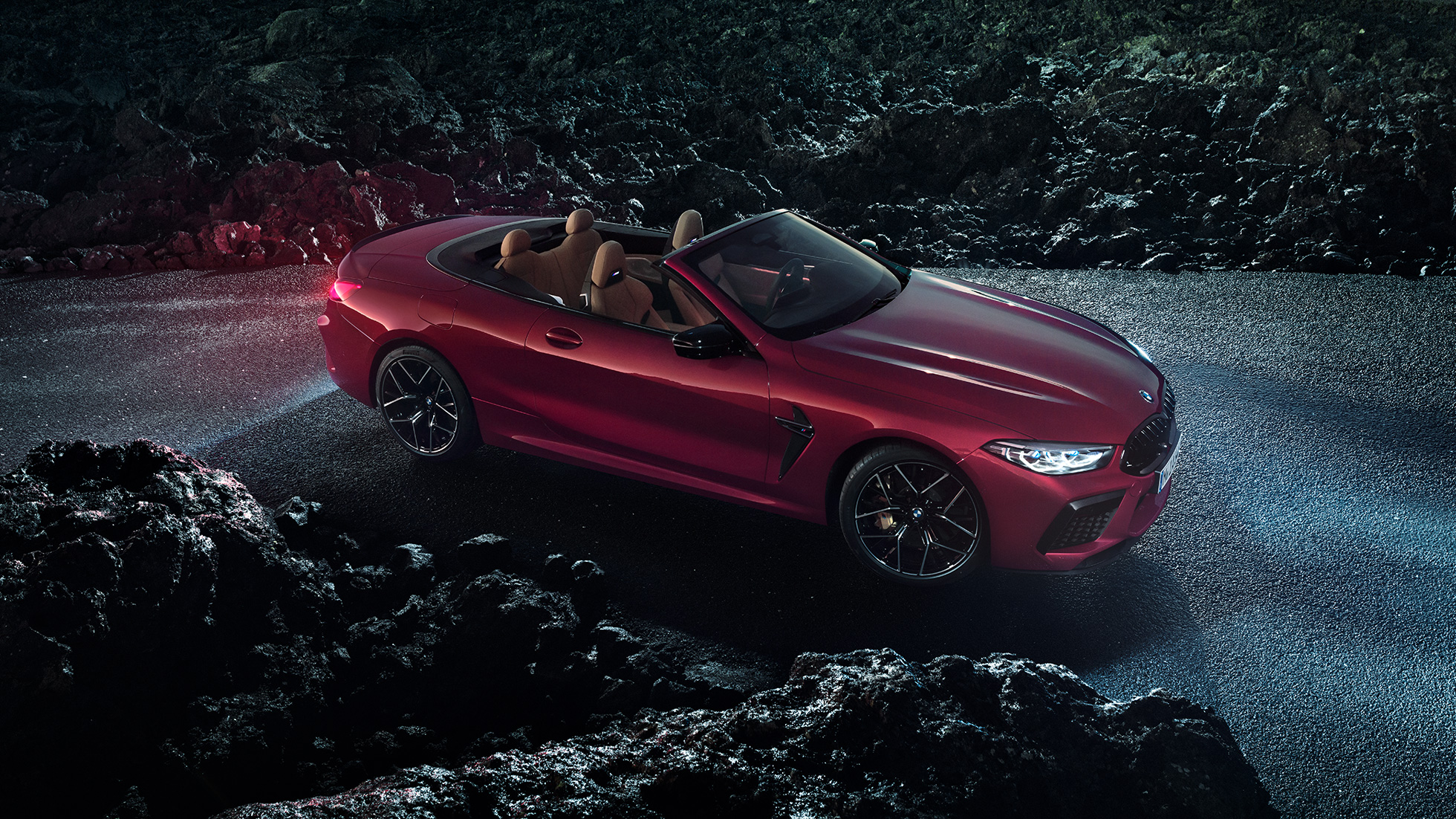 BMW M8 Competition Cabrio, vista lateral a três quartos. BMW M8 Competition Cabrio na cor exterior vermelho Motegi metalizada com a capota flexível aberta, parado no asfalto, à noite, vista lateral a três quartos.