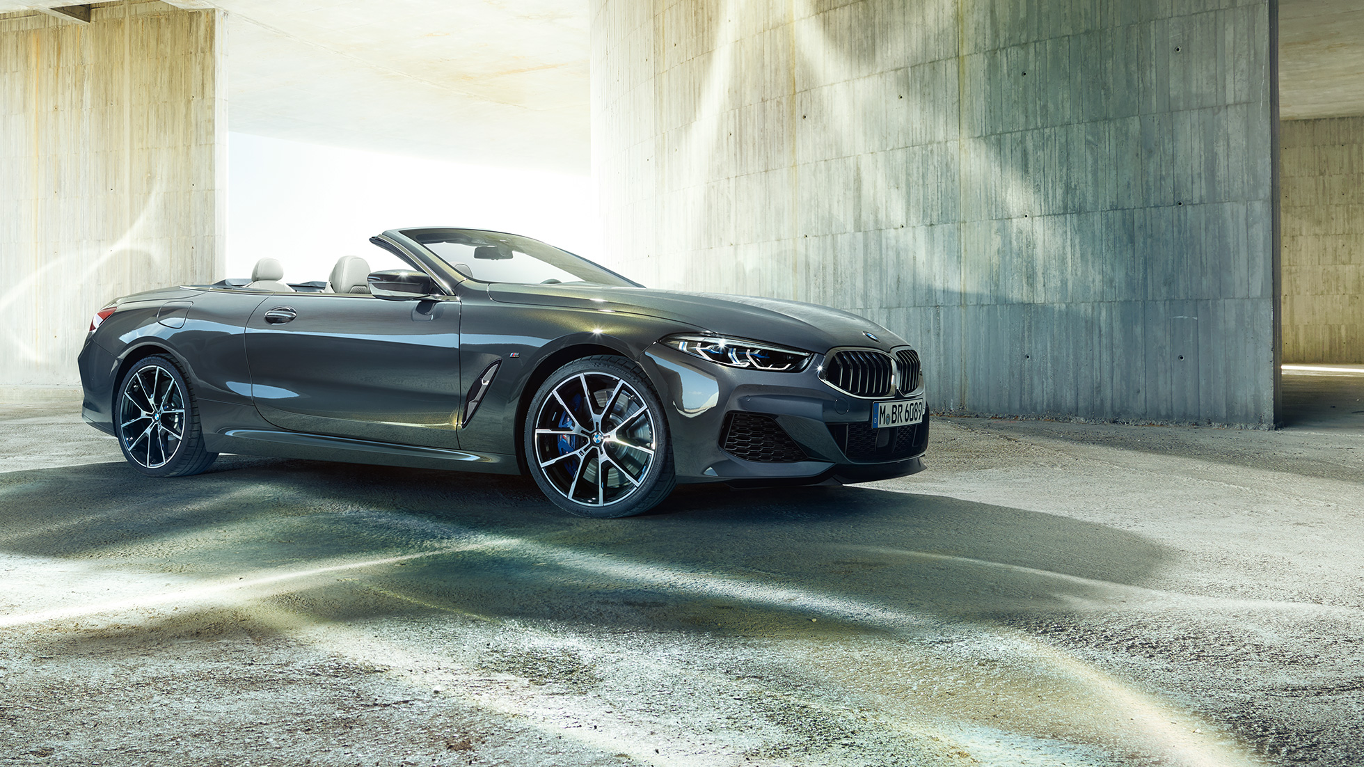 BMW M850i xDrive Cabrio, perspetiva lateral a três quartos. BMW M850i xDrive Cabrio na pintura cinza Dravit metalizada com capota flexível aberta, em frente a um edifício em betão, perspetiva lateral a três quartos.