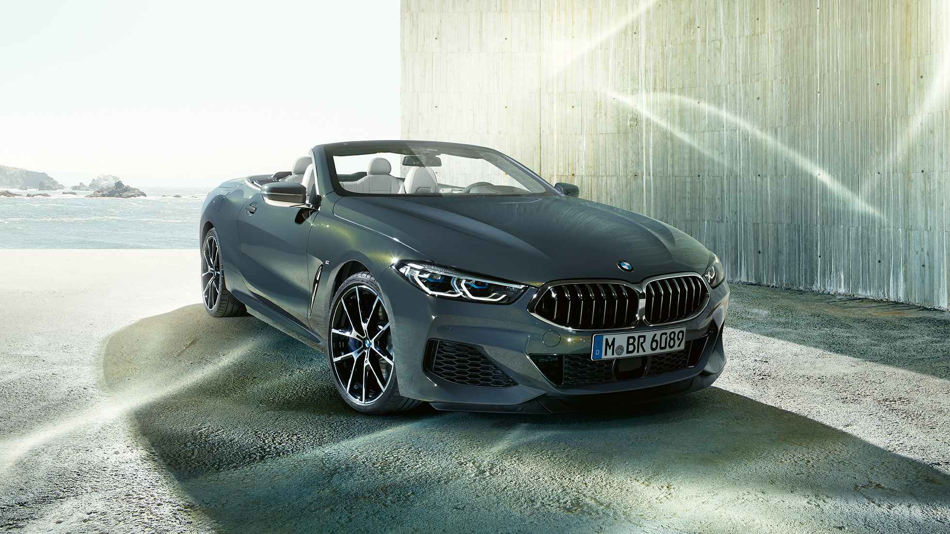 BMW M850i xDrive Cabrio, perspetiva frontal a três quartos. BMW M850i xDrive Cabrio na pintura cinza Dravit metalizada com capota flexível aberta, em frente ao mar, perspetiva frontal a três quartos.