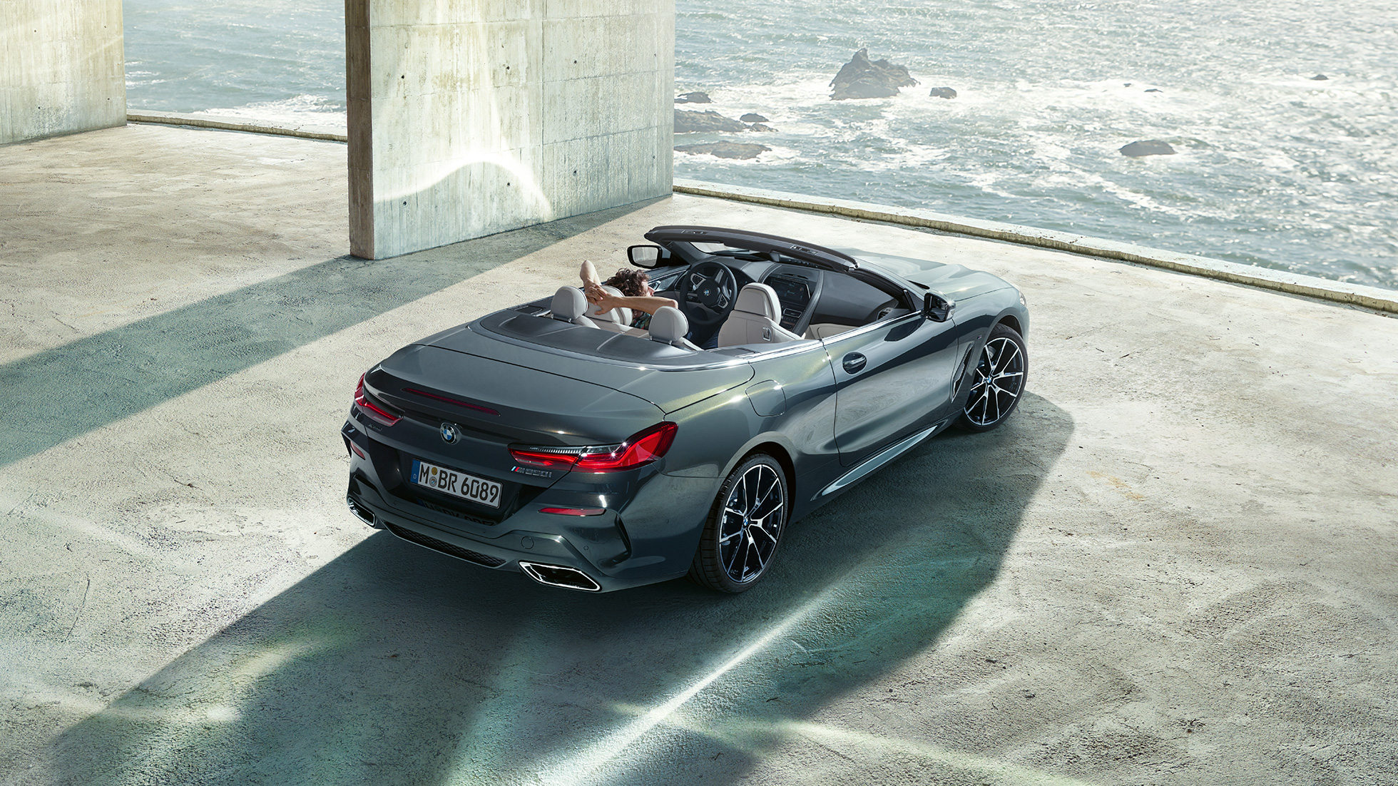 BMW M850i xDrive Cabrio, perspetiva aérea. BMW M850i xDrive Cabrio na pintura cinza Dravit metalizada com capota flexível aberta, estacionado no interior de um edifício em betão, em frente ao mar, perspetiva aérea.