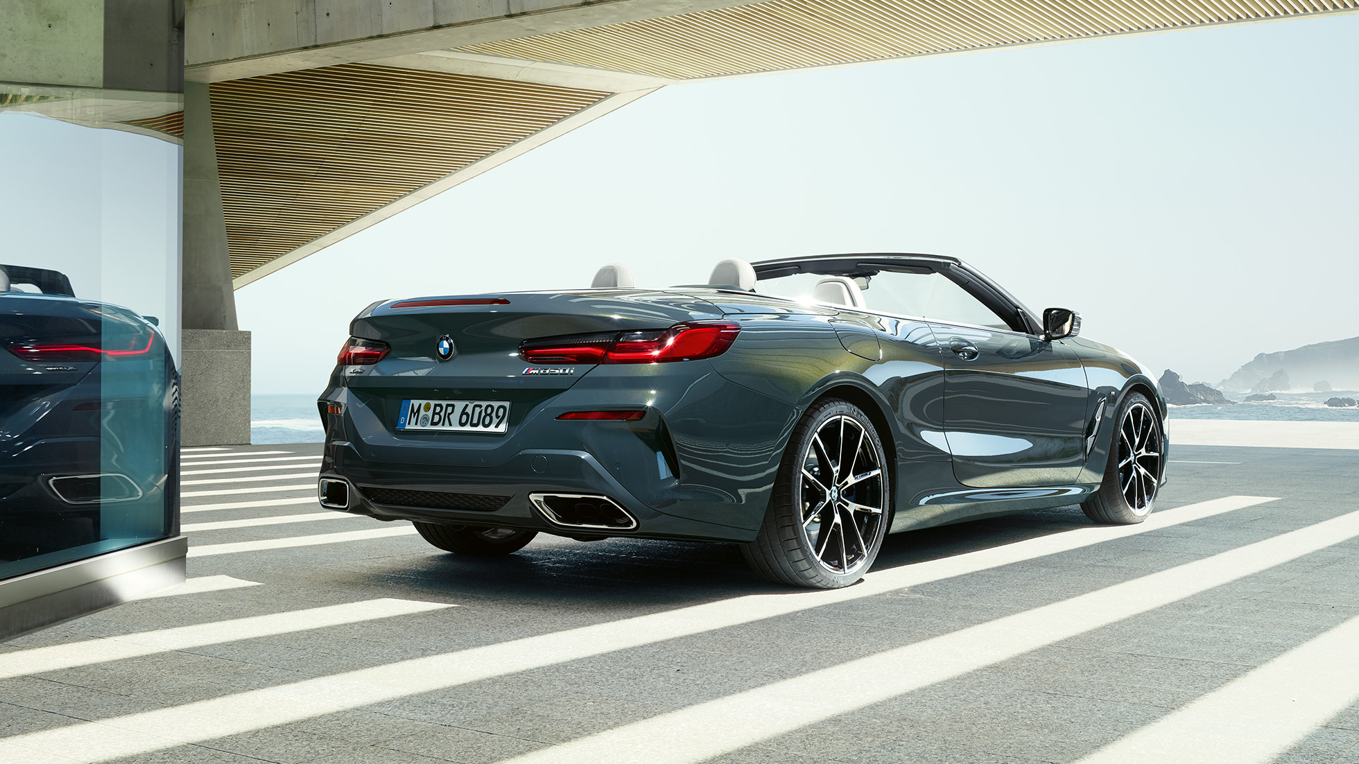 BMW M850i xDrive Cabrio, perspetiva traseira a três quartos. BMW M850i xDrive Cabrio na pintura cinza Dravit metalizada com capota flexível aberta, a estacionar num local com perspetiva para o mar, perspetiva traseira a três quartos.