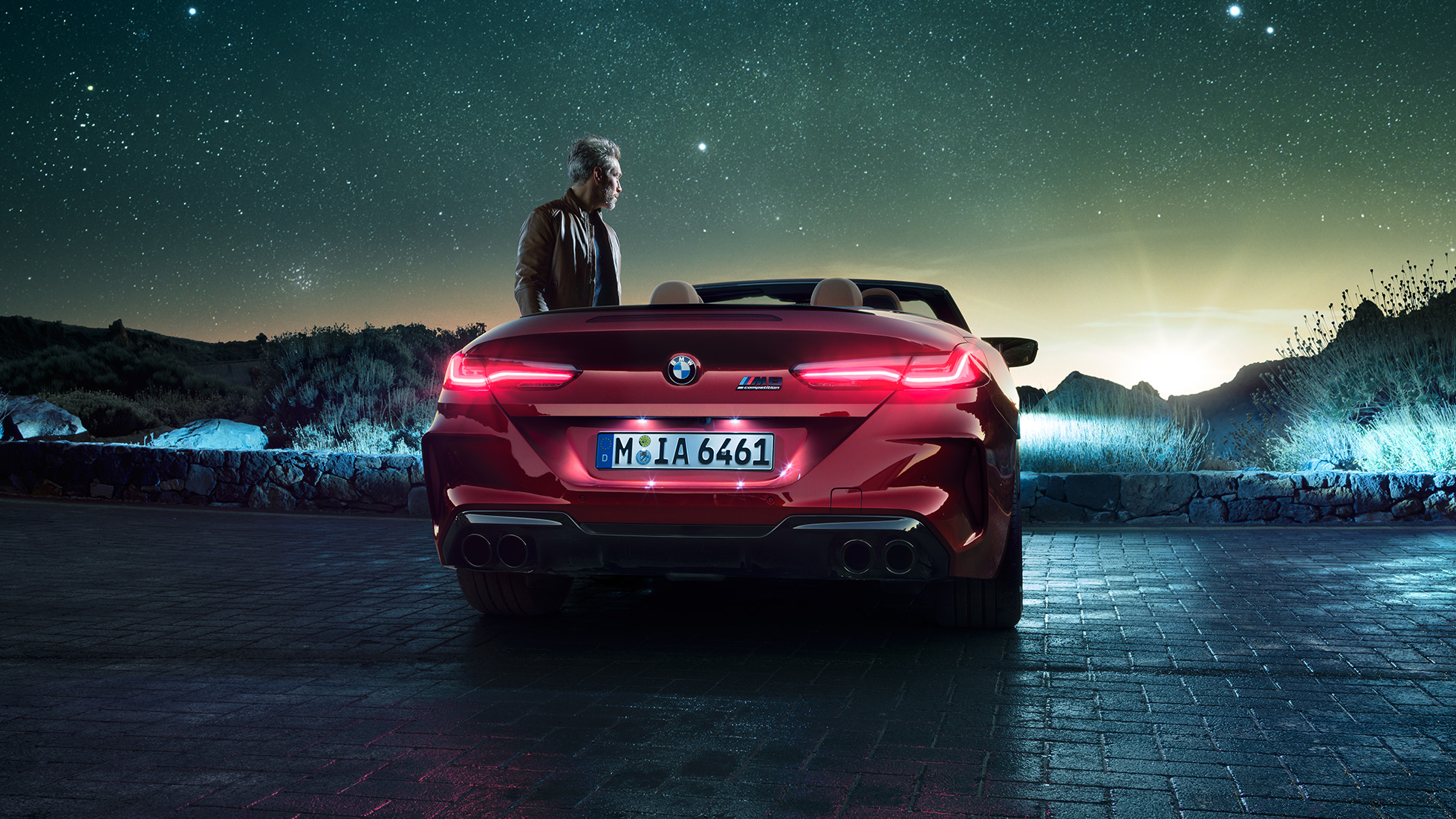 BMW M8 Competition Cabrio, perspetiva traseira. BMW M8 Competition Cabrio na pintura vermelho Motegi metalizada com capota flexível aberta, parado, com um homem sob um céu estrelado, perspetiva traseira.