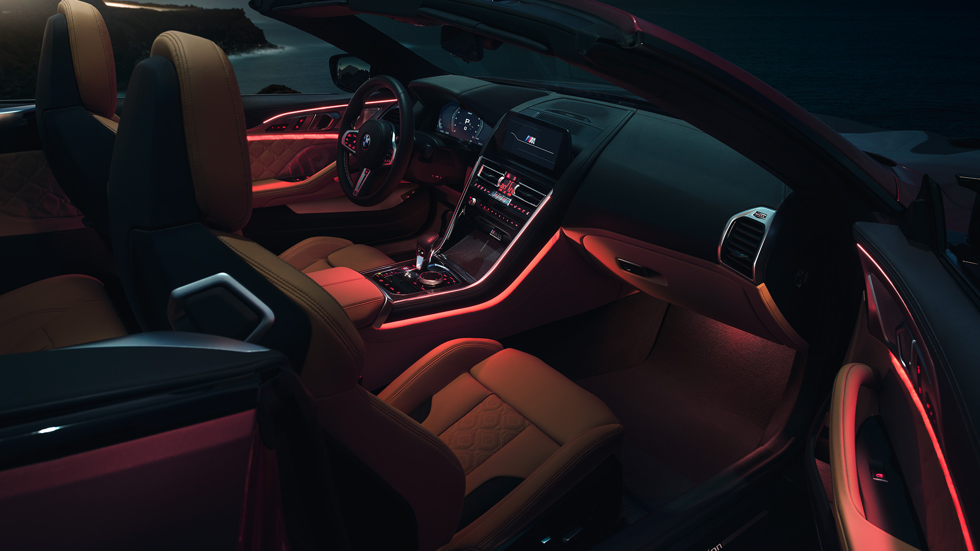 BMW M8 Competition Cabrio, interior. BMW M8 Competition Cabrio com capota fexível aberta, interior com luz ambiente em vermelho, à noite.