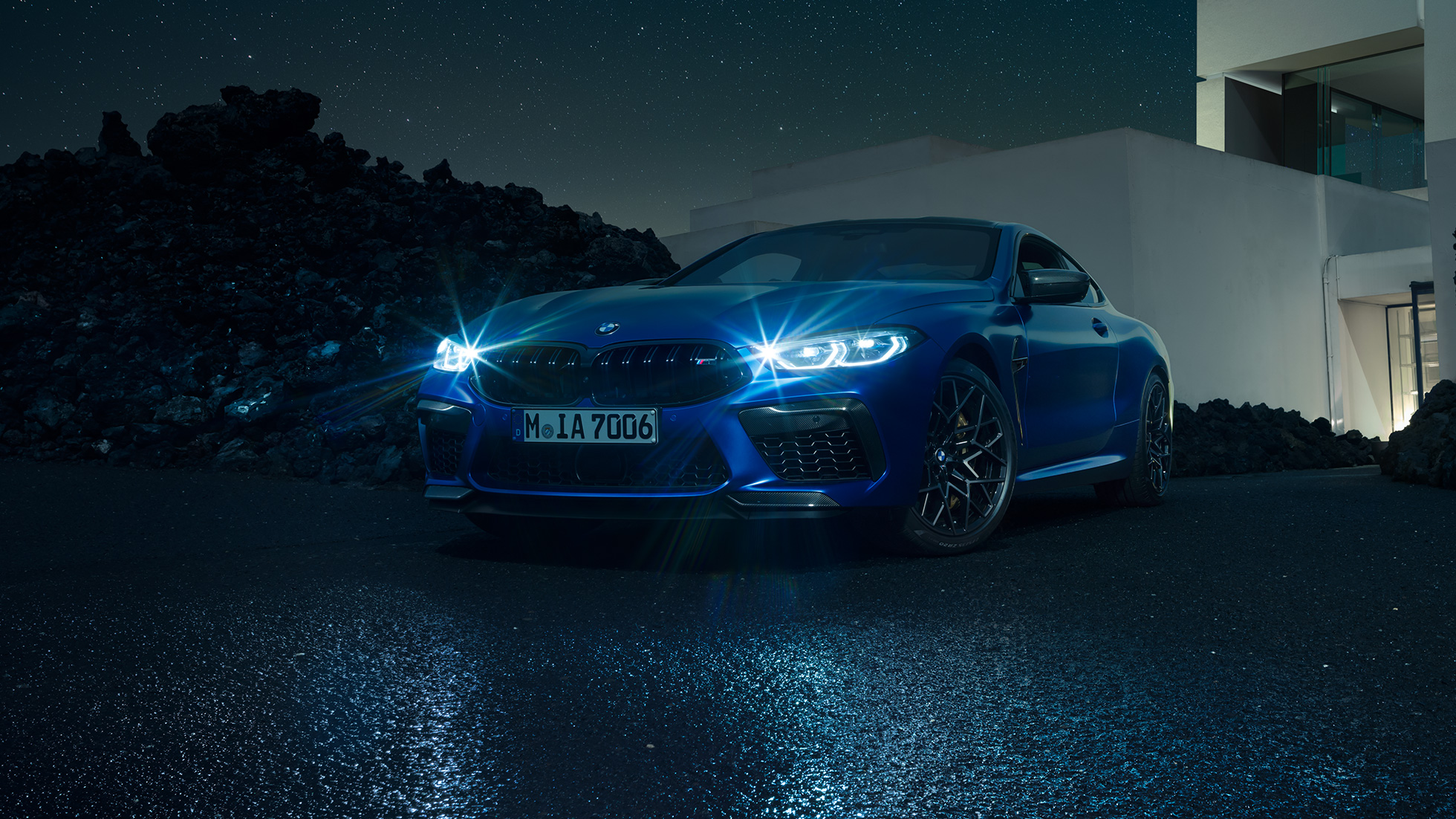 BMW M8 Competition Coupé, perspetiva frontal a três quartos. BMW M8 Competition Coupé na pintura azul Marina Bay metalizada com faróis LED iluminados, parado em frente a uma moradia, à noite, perspetiva frontal a três quartos.
