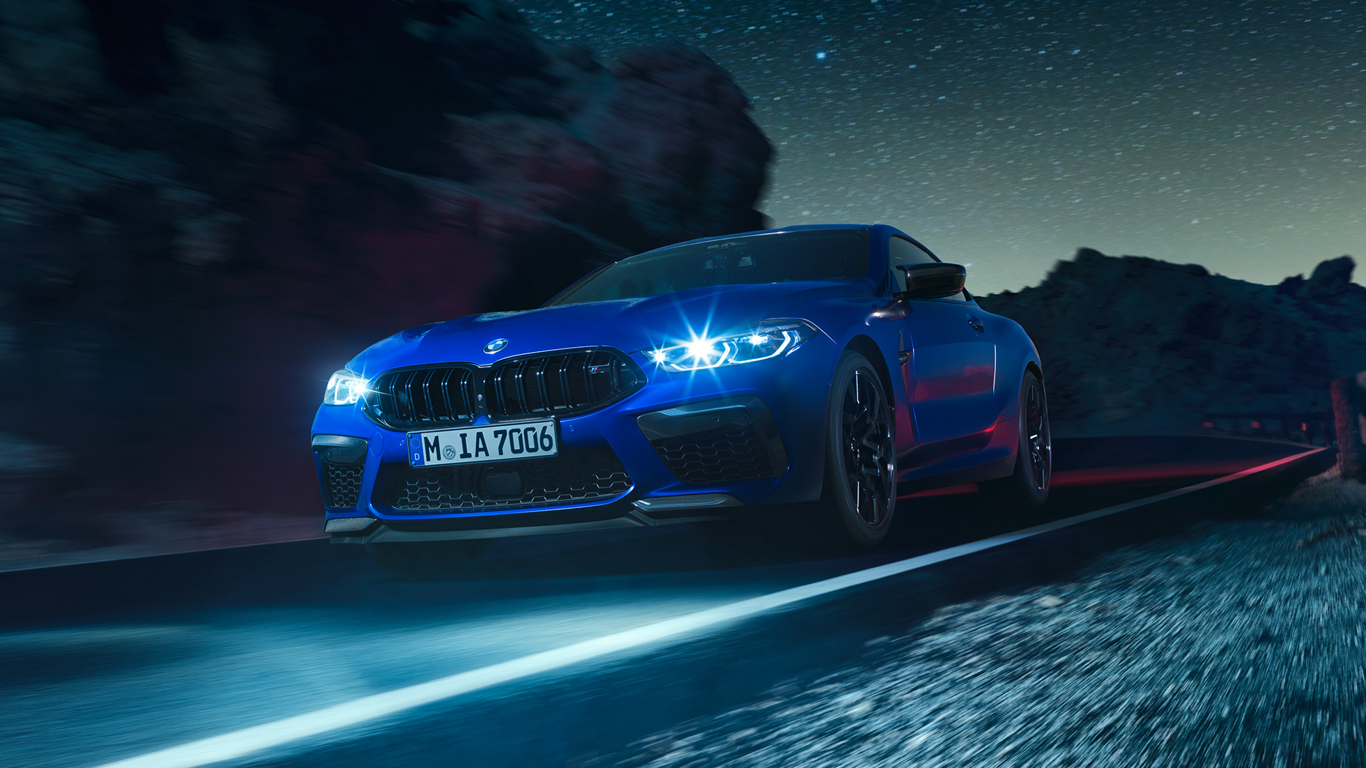 BMW M8 Competition Coupé em movimento, perspetiva frontal a três quartos. BMW M8 Competition Coupé na pintura azul Marina Bay metalizada com faróis LED iluminados, em movimento, à noite, perspetiva frontal a três quartos.