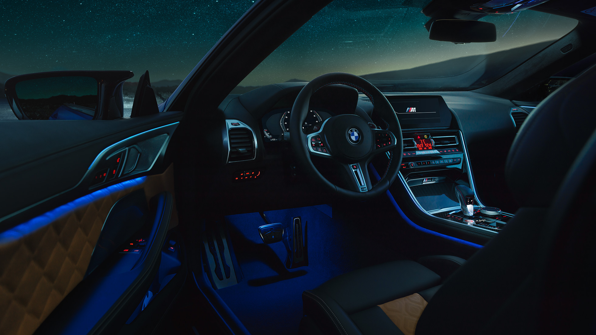 Interior do BMW M8 Competition Coupé. BMW M8 Competition Coupé, interior à noite, com volante M em pele e luz ambiente em azul, com a porta do condutor aberta.